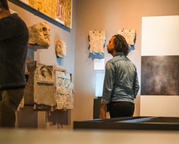 Young Caucasian woman reading through a digital tablet with data of the displayed objects while her biracial husband is checking out the stone remains of an Ancient Roman town. Candid shot over the display cases in the middle of museum gallery.